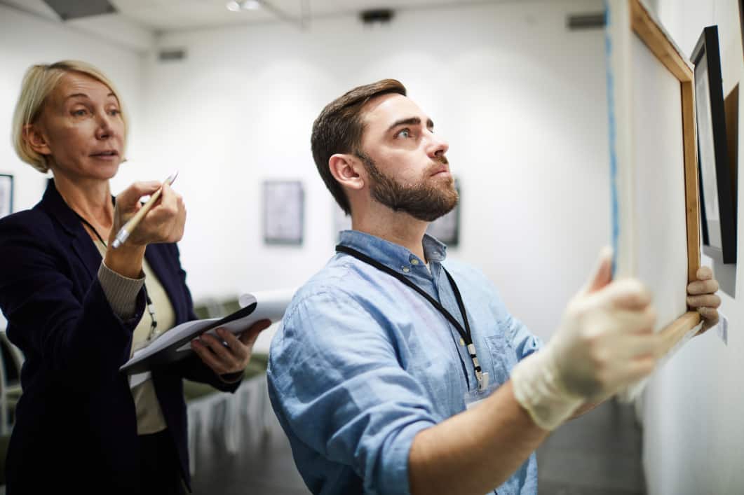 Man hangs art in gallery while woman holding clipboard asks how to authenticate art