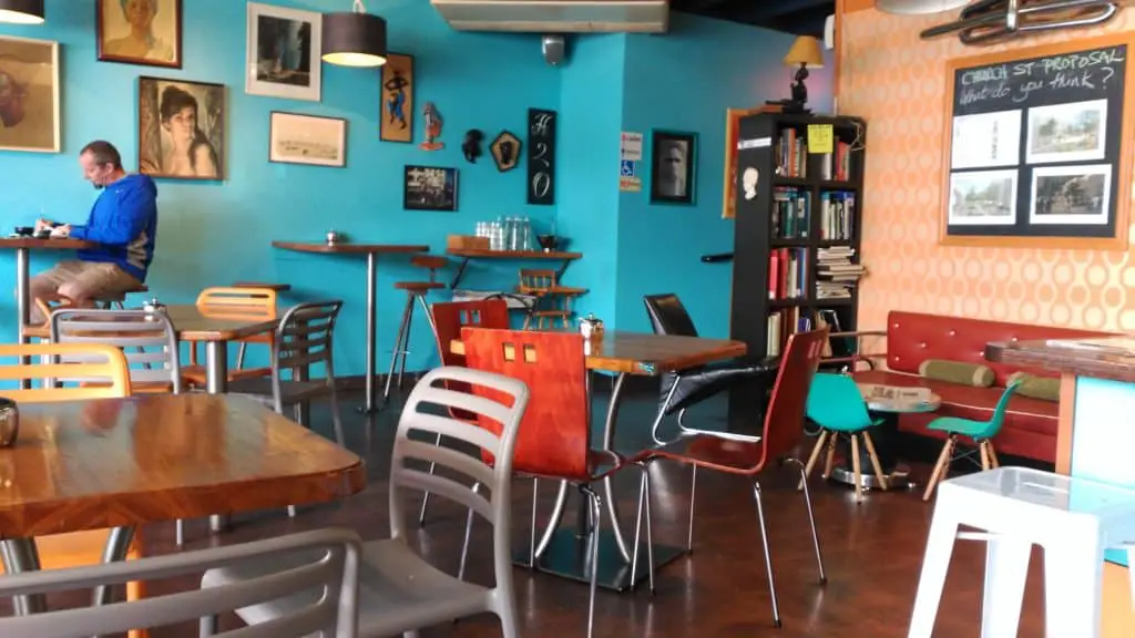 Man sitting in coffee shop with art from emerging artists hanging on wall behind him.