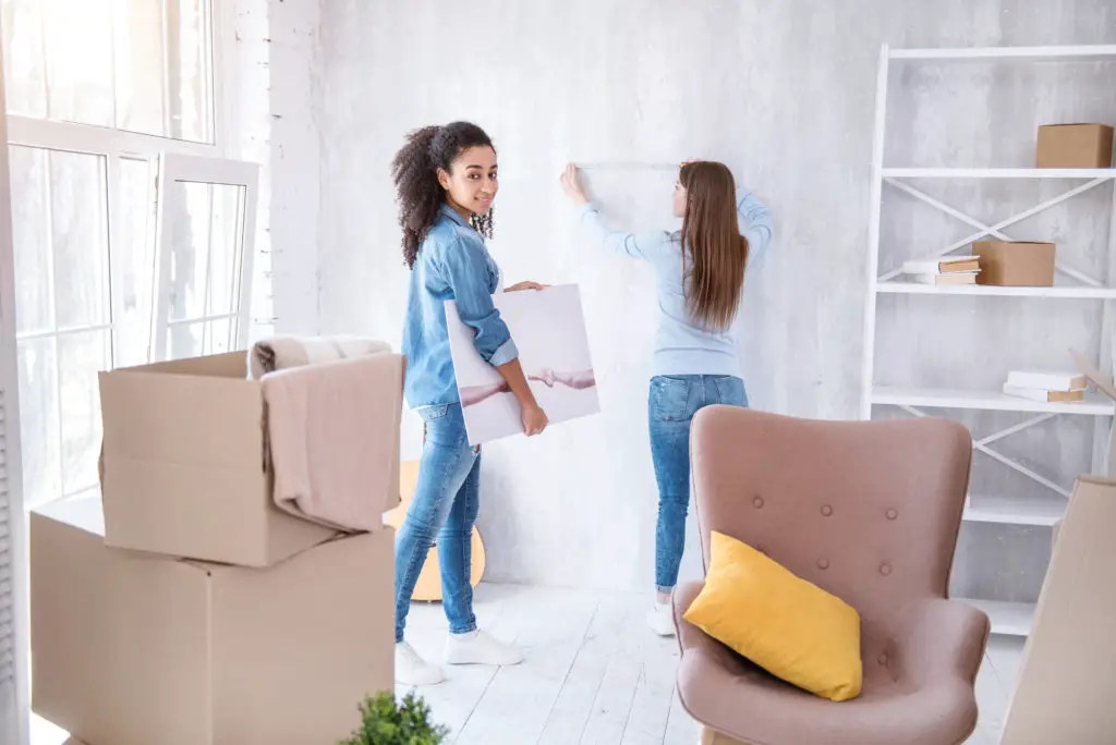 One woman holds a canvas print while another attaches adhesive tape to wall