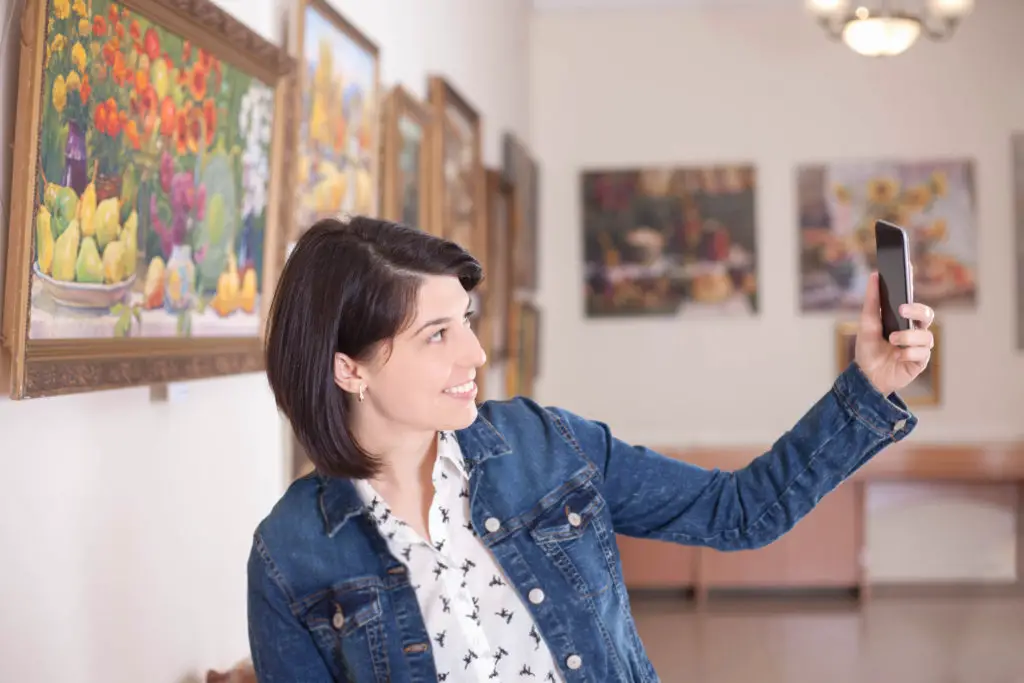 Woman in a gallery taking a selfie making sure she's in the phonograph with the painting