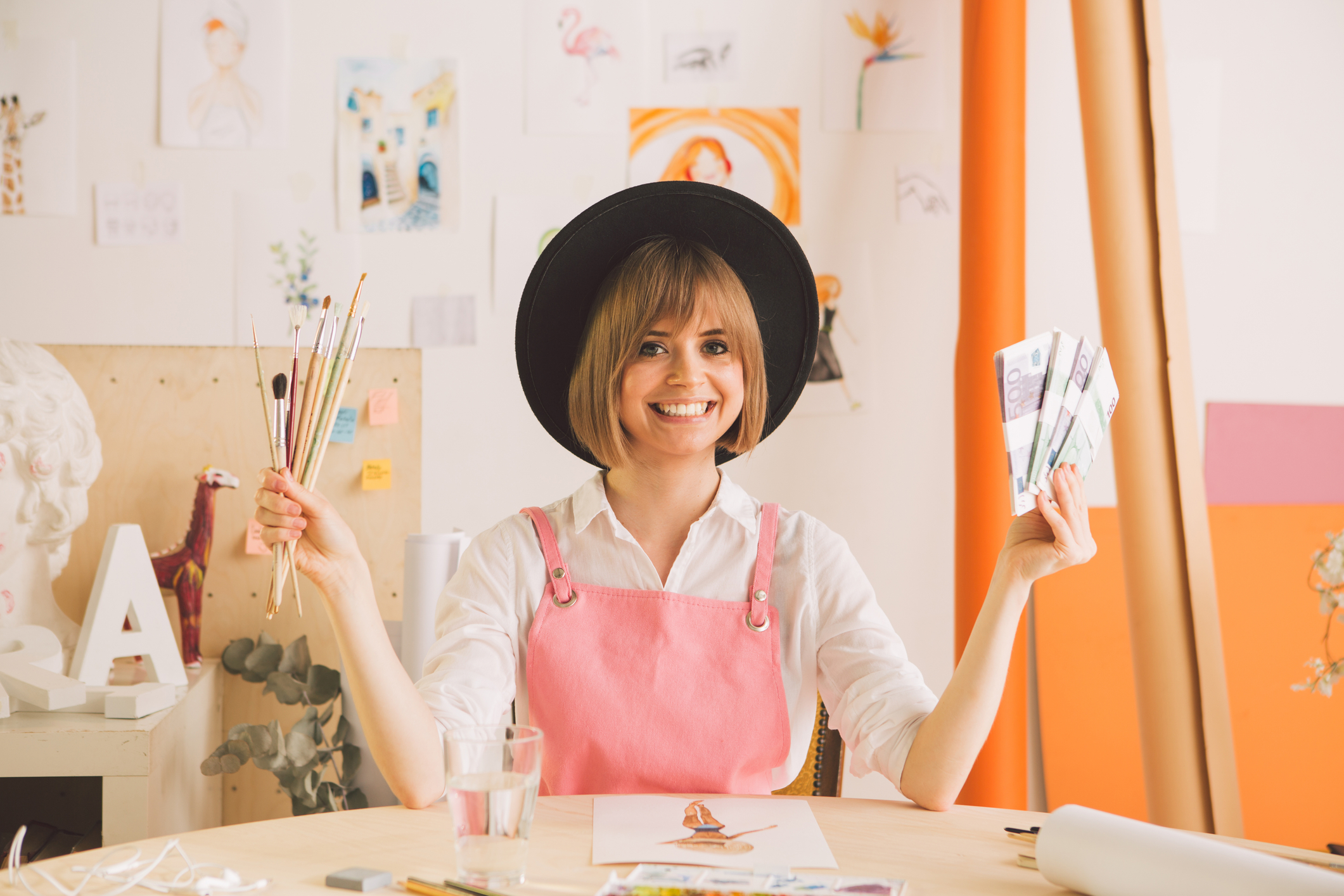 Female artist wearing a hat holding paintbrushes and money.