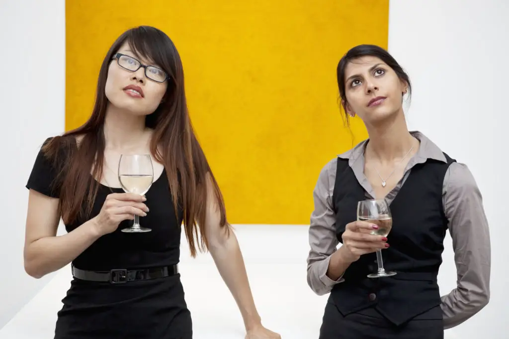 Two women holding wine glasses at a gallery exercising good art etiquette