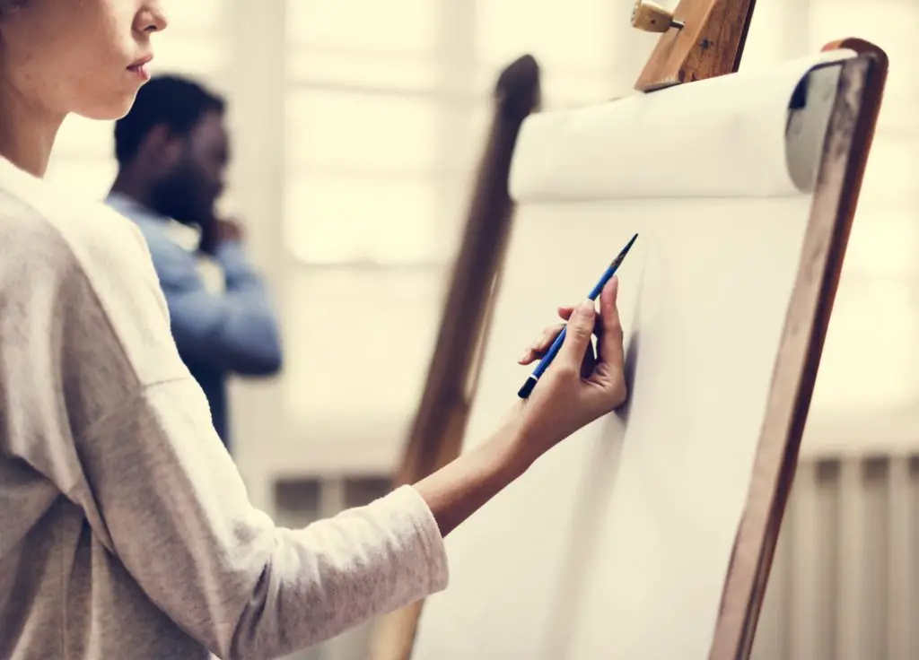 Woman holding a pencil in art class in front of an easel deciding between drawing vs painting