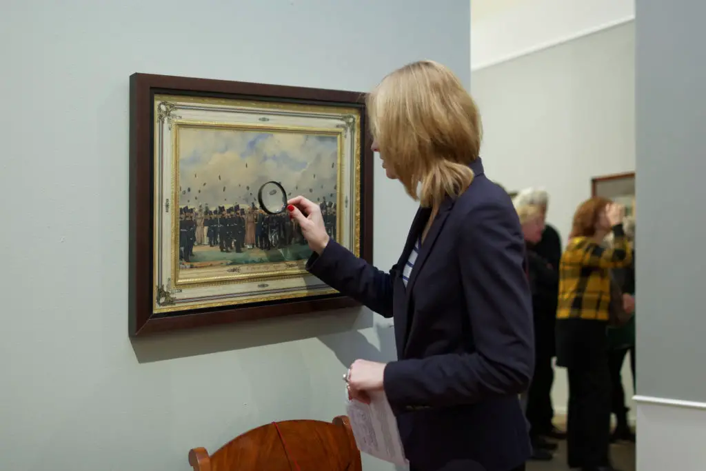 Woman using a magnifying glass to appraise a painting on the wall.