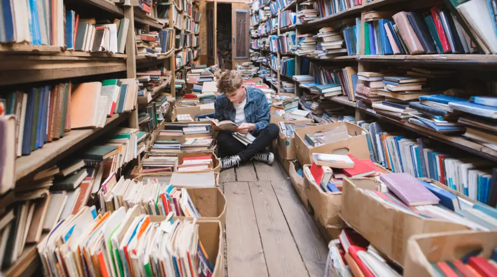 Man sitting on the floor among books in a library isle researching an artwork's history.