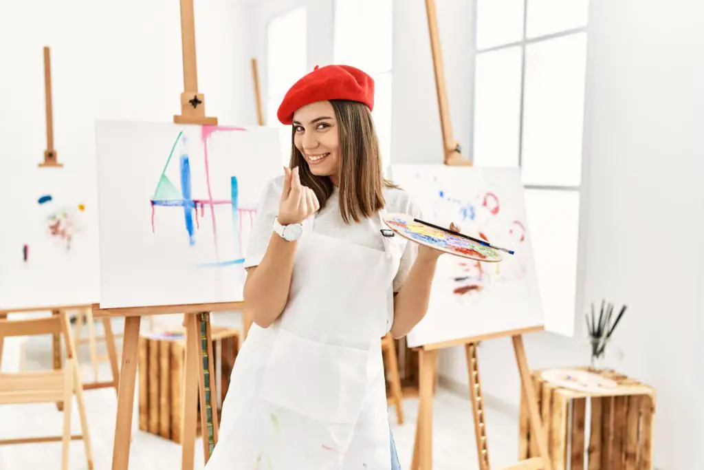 Female artist wearing a red beret in front of easel making the money gesture with her fingers.