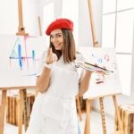 Female artist wearing a red beret in front of easel making the money gesture with her fingers.