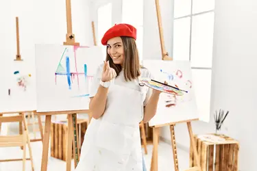 Female artist wearing a red beret in front of easel making the money gesture with her fingers.