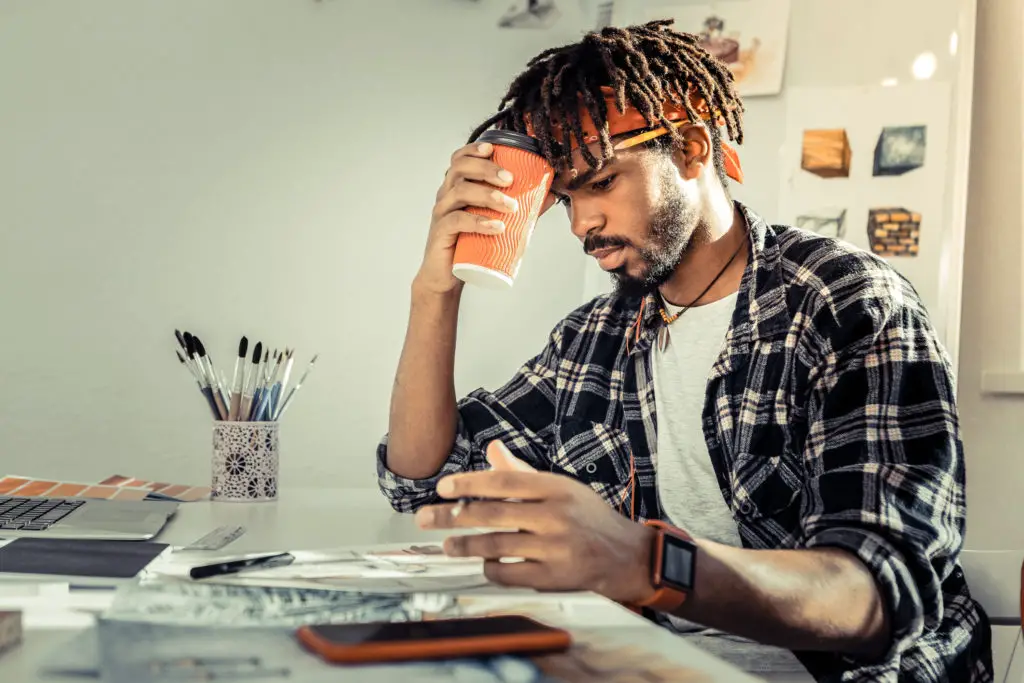 Male artist calculates his expenses while holding a coffee cup to compute how much he should charge for his art.