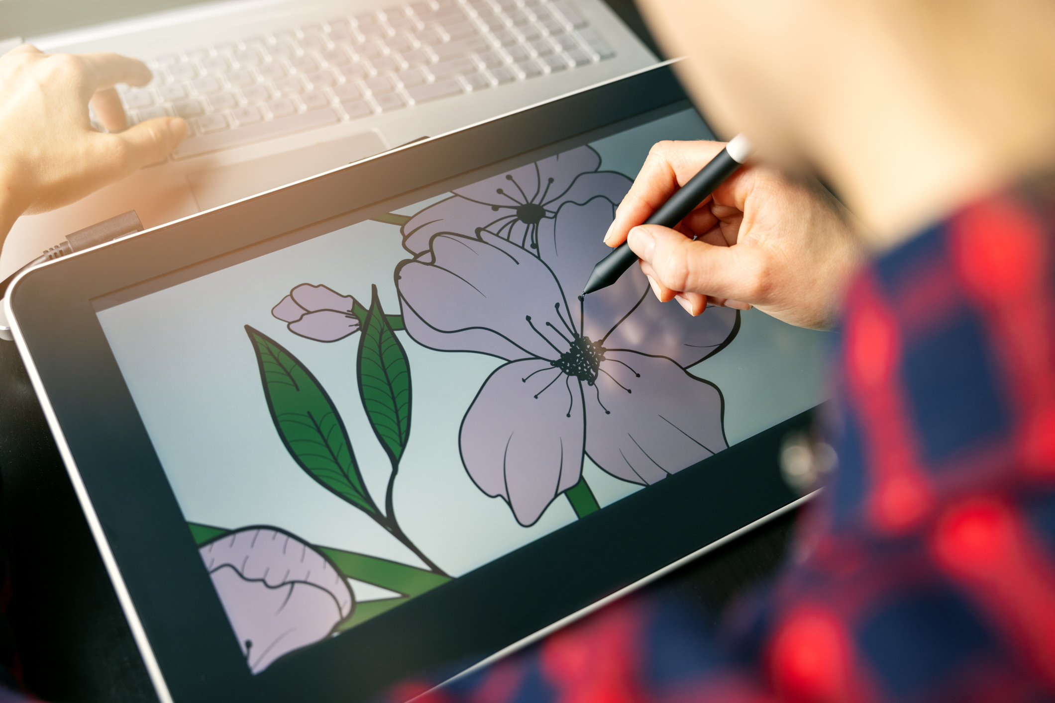 Over the shoulder of a male digital artist as he sketches flowers on a digital sketch pad hooked up to his computer.