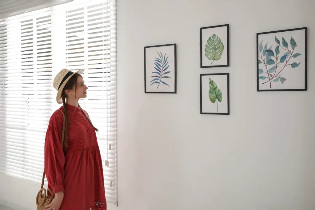 Woman in a hat and braids looking at artist reproduction prints of different leaves hanging on a wall.